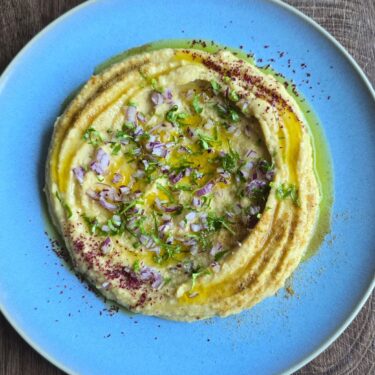 Santorini auf dem Teller: Fava – cremig, sonnig, mediterran 4 Angerichtetes Fava auf blauem Teller mit Olivenöl, Petersilie und roten Zwiebeln.