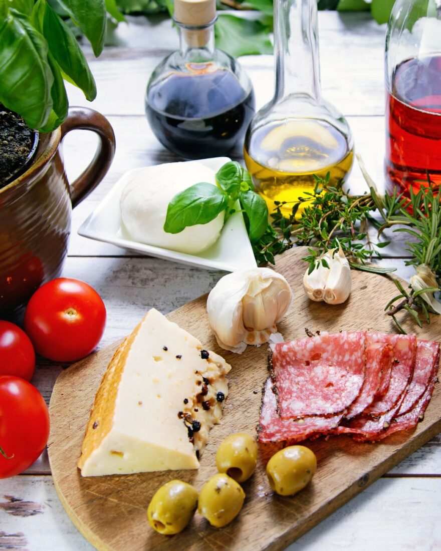 Nachhaltige Gastronomie mit Wirkung: Herkunft entscheidet 2 Mediterrane Zutaten auf einem Holztisch mit Olivenöl, Käse, Wurst und frischen Kräutern