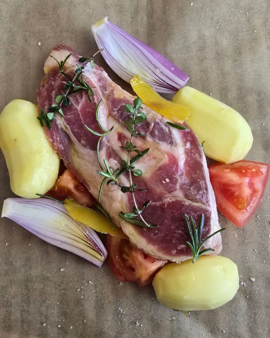 Eine rohe Lammscheibe mit Kartoffeln, Tomaten, Schalotten und Kräutern auf Backpapier arrangiert