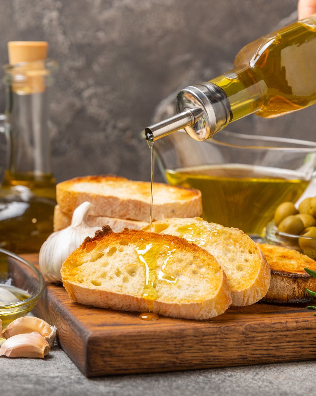 Geröstete Brotscheiben mit goldgrünem Olivenöl, das frisch aus der Flasche gegossen wird. Im Hintergrund Knoblauch, Oliven und Glasflasche mit Öl.