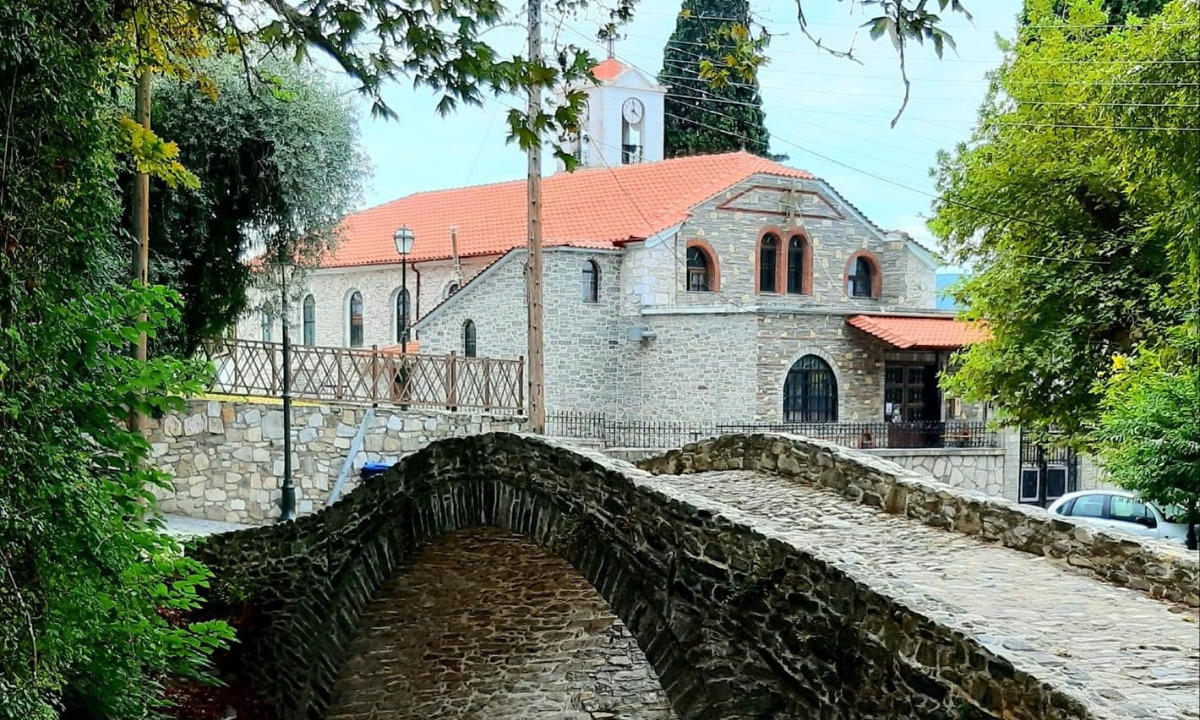 Das Bild zeigt im Hintergrund die Kirche im Dorf Moustheni mit rotem Dach und Steinmauer. Neben der Kirche erkennt man einen Holzzaun und im Vordergrund sieht man eine typische Brücke mit einem Bogen aus Stein gemauert.