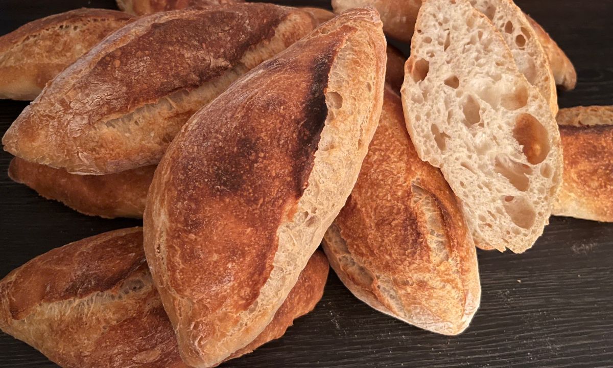 Auf dem vorliegenden Bild sehen wir ein ansprechendes kulinarisches Arrangement, das die Kunst des Brotbackens zelebriert. Ein duftendes Baguette, zubereitet mit Liebe und traditionellen Techniken, ist das Herzstück dieser Darstellung. Das Baguette liegt auf einer dunklen Arbeitsplatte mit Holzmasserung. Die goldene, knusprige Kruste des Baguettes kontrastiert wunderbar mit dem dunklen Holz des Brettes und lässt das Brot umso einladender wirken. Die Oberfläche des Baguettes ist von feinen Rissen und Blasen gezeichnet, ein Zeichen für die perfekte Brottextur, die beim Backen erreicht wurde. Dieses Baguette wurde mit Liebe und Geduld gebacken und enthüllt seine innere Pracht, sobald es aufgeschnitten wird. Die Krume des Baguettes zeigt eine herrliche Mischung aus Luftigkeit und Dichte, was auf die Verwendung von Lievito Madre, einem natürlichen Sauerteig, hinweist. Die unverkennbare Textur und der köstliche Geschmack, der durch diesen traditionellen Sauerteig entsteht, verleihen dem Baguette eine besondere Note. Insgesamt präsentiert das Bild eine Hommage an die Kunst des Brotbackens und lädt den Betrachter dazu ein, die einfache, aber wunderbare Freude eines frisch gebackenen Baguettes zu genießen. Es weckt die Vorstellung von Geschmack und Textur, die dieses Brot zu einem zeitlosen kulinarischen Genuss machen.